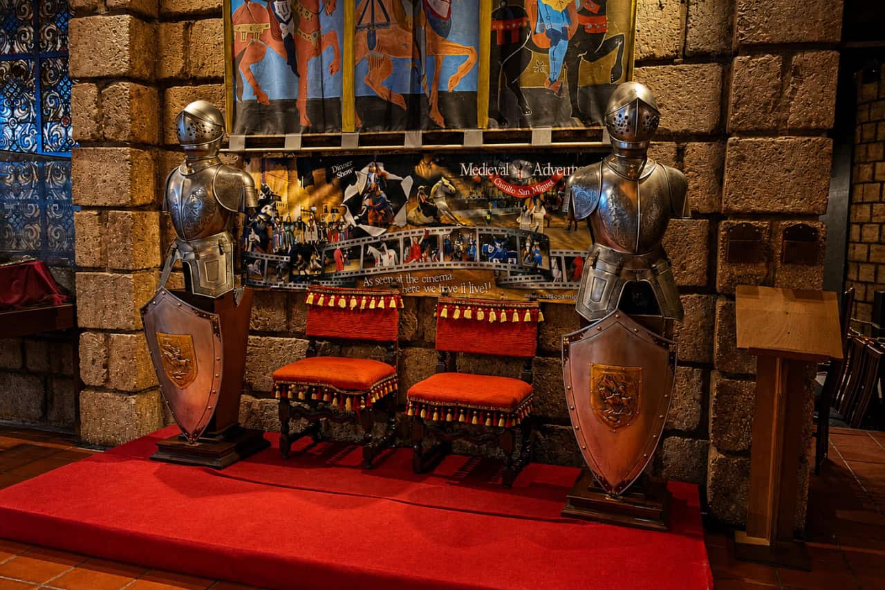 Two suits of armor with shields and red chairs inside the Medieval show Castillo San Miguel venue.