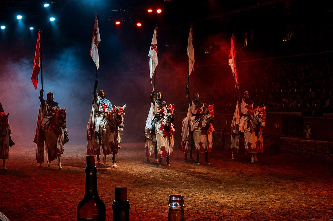 Mounted knights hold flags under red and blue lights during the medieval show San Miguel de Abona arena scene.