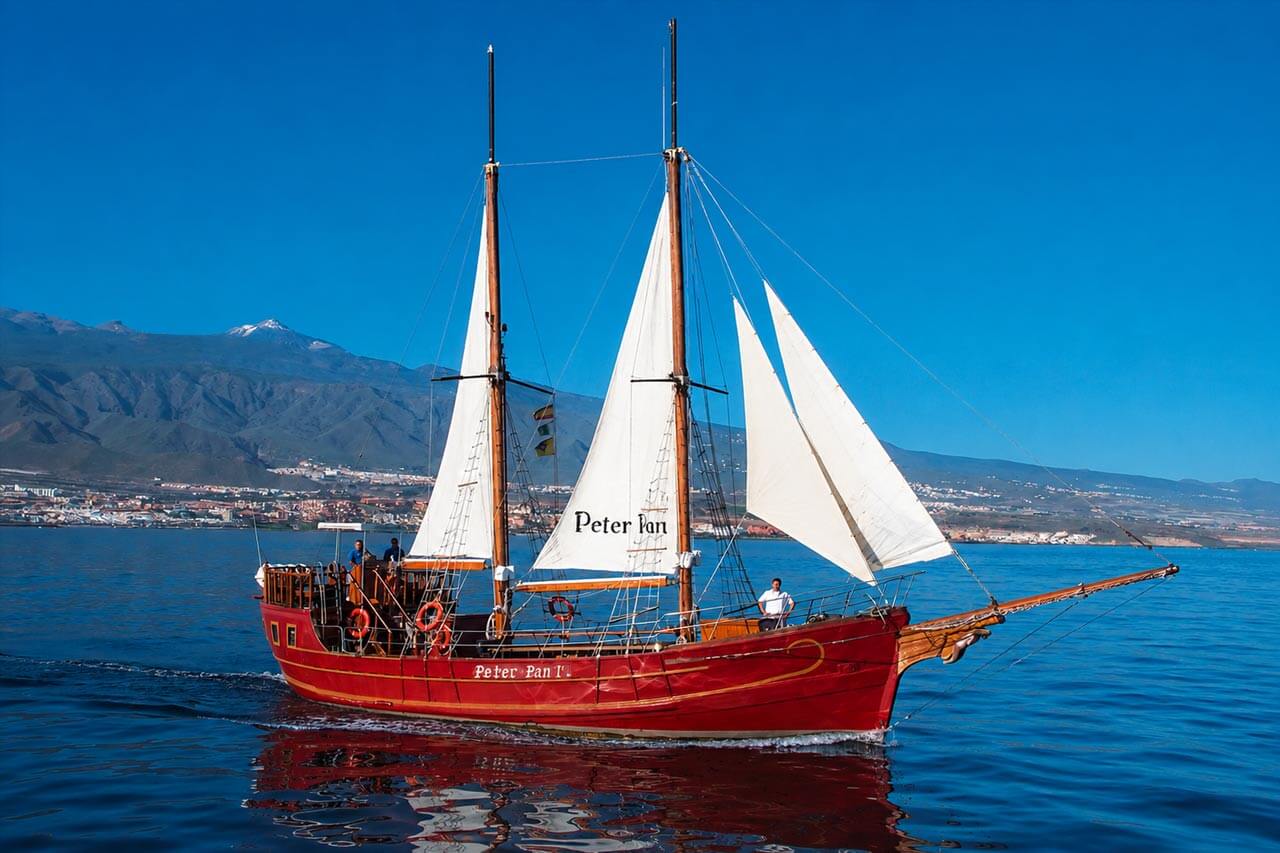 The red Peter Pan sailing boat cruising across the Atlantic Ocean near Tenerife with its white sails raised, guests on deck and the island’s volcanic mountains in the background.