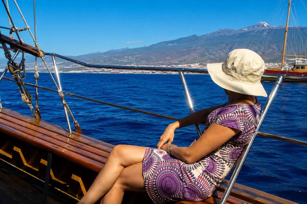 A guest relaxing on the polished wooden side deck of the Peter Pan sailing boat while looking across the Atlantic Ocean toward Tenerife’s volcanic coastline.
