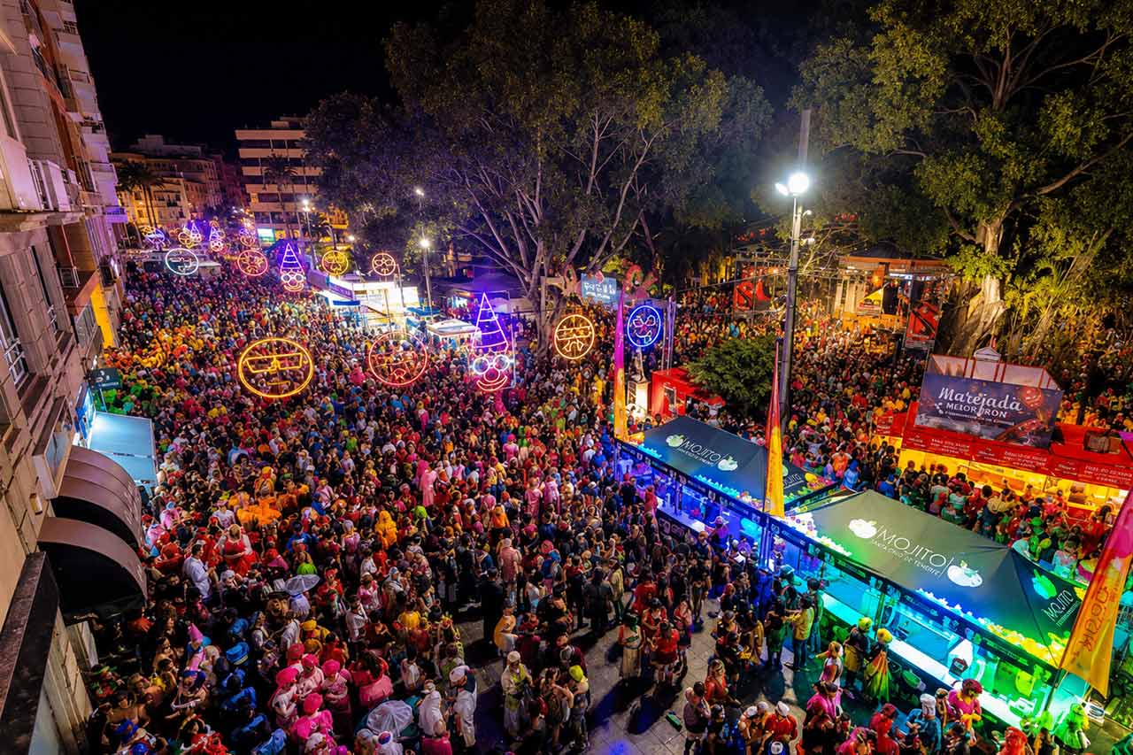 Panoramiczny nocny widok na uliczne obchody karnawału w Santa Cruz de Tenerife, z tysiącami ludzi wypełniającymi centrum miasta pod kolorowymi, oświetlonymi dekoracjami. Jasne bary festiwalowe, drzewa, oświetlenie uliczne i tłumy w kostiumach ukazują energiczną atmosferę nocnego życia, jakiej odwiedzający mogą doświadczyć podczas karnawału.