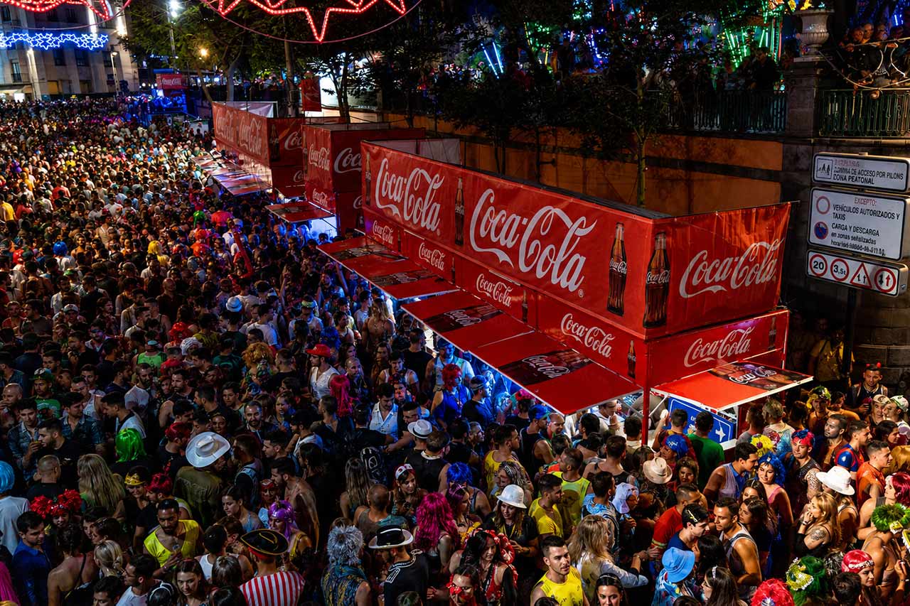 Una concurrida escena nocturna en el Carnaval de Santa Cruz de Tenerife, con miles de visitantes disfrazados reunidos alrededor de puestos de comida y bebida brillantemente iluminados. Grandes puestos de refrescos de la marca Coca-Cola se encuentran en el centro de la multitud, rodeados de coloridas decoraciones de carnaval, iluminación callejera y personas con disfraces elaborados, capturando la animada vida nocturna y el ambiente festivo en el centro de la ciudad.