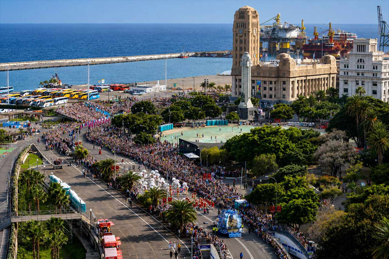 Vista panorámica de la ruta del desfile del Carnaval de Santa Cruz de Tenerife cerca del paseo marítimo. Miles de espectadores abarrotan las calles mientras coloridos artistas y agrupaciones carnavaleras avanzan por la ciudad, con la Plaza de España, edificios históricos, el puerto, palmeras y el océano Atlántico visibles al fondo