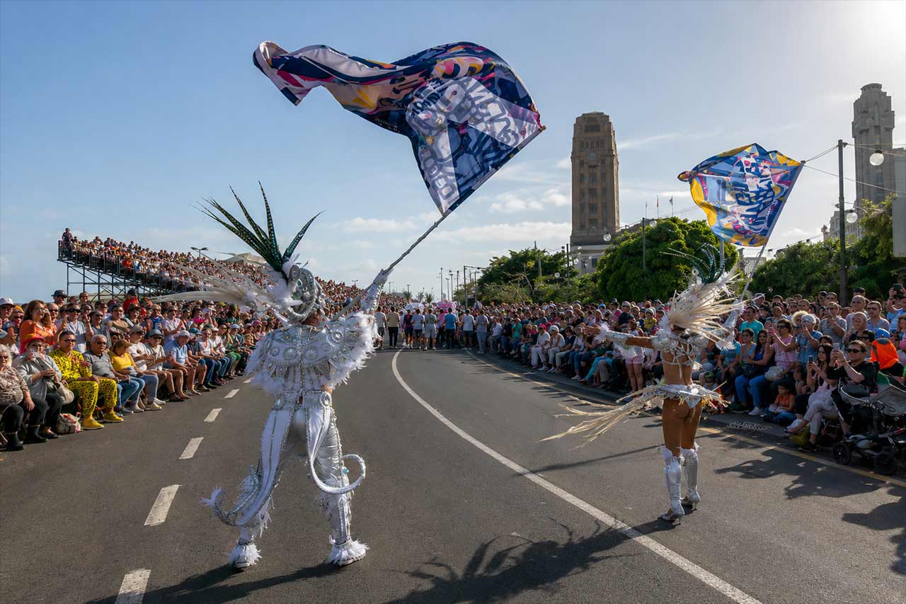 Artistas disfrazados del carnaval agitaban grandes banderas de colores mientras avanzaban por una ruta de desfile abarrotada en Santa Cruz de Tenerife. Los espectadores llenaban ambos lados de la calle mientras los artistas, vestidos con elaborados trajes, aportaban movimiento, color y espectáculo a uno de los principales desfiles diurnos del Carnaval de Tenerife.