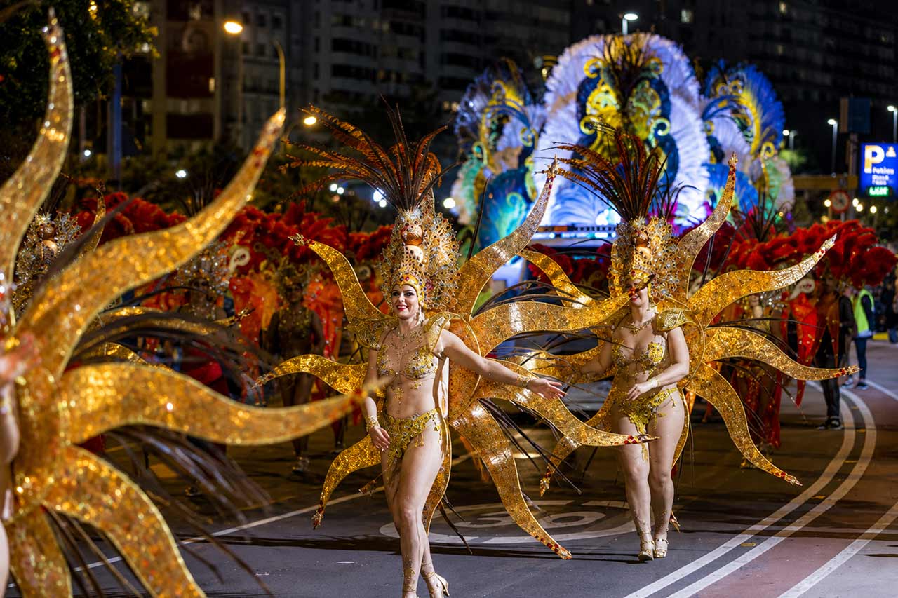 Los artistas del carnaval con espectaculares trajes dorados en forma de sol participan en un desfile nocturno por Santa Cruz de Tenerife. La brillante iluminación de las calles, los trajes de plumas rojas y una gran pantalla de carnaval iluminada en el fondo crean la atmósfera dramática asociada con las celebraciones del carnaval nocturno de Tenerife.