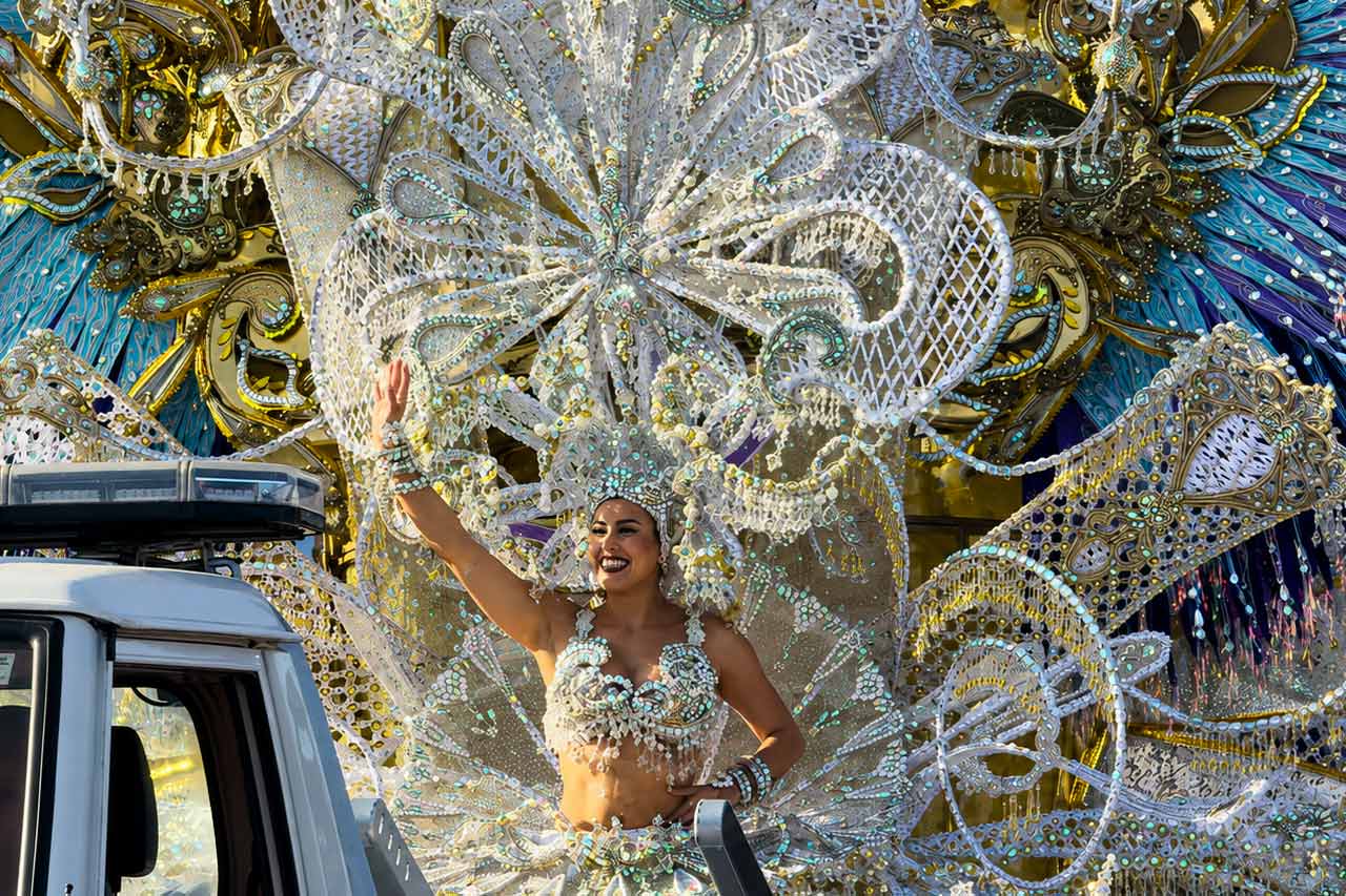 Una artista de Reina del Carnaval saluda desde una elaborada carroza durante el Carnaval de Santa Cruz de Tenerife. Está rodeada por un espectacular despliegue de vestuario en plata, oro y turquesa, cubierto de intrincadas decoraciones, lentejuelas y detalles escultóricos. La imagen captura la escala, la artesanía y el espectáculo visual de las carrozas de carnaval y las actuaciones de las reinas, que son uno de los principales atractivos de las famosas celebraciones anuales de Tenerife.