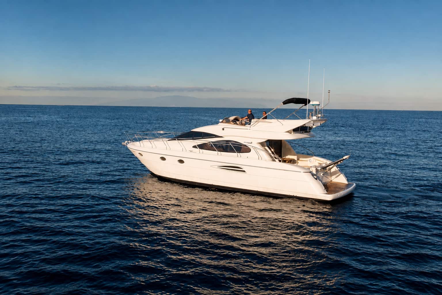 The white Seaduction motor yacht cruising on calm Atlantic water near Tenerife, with guests seated on the upper deck and clear views of the yacht’s spacious exterior, shaded flybridge and rear swimming platform.