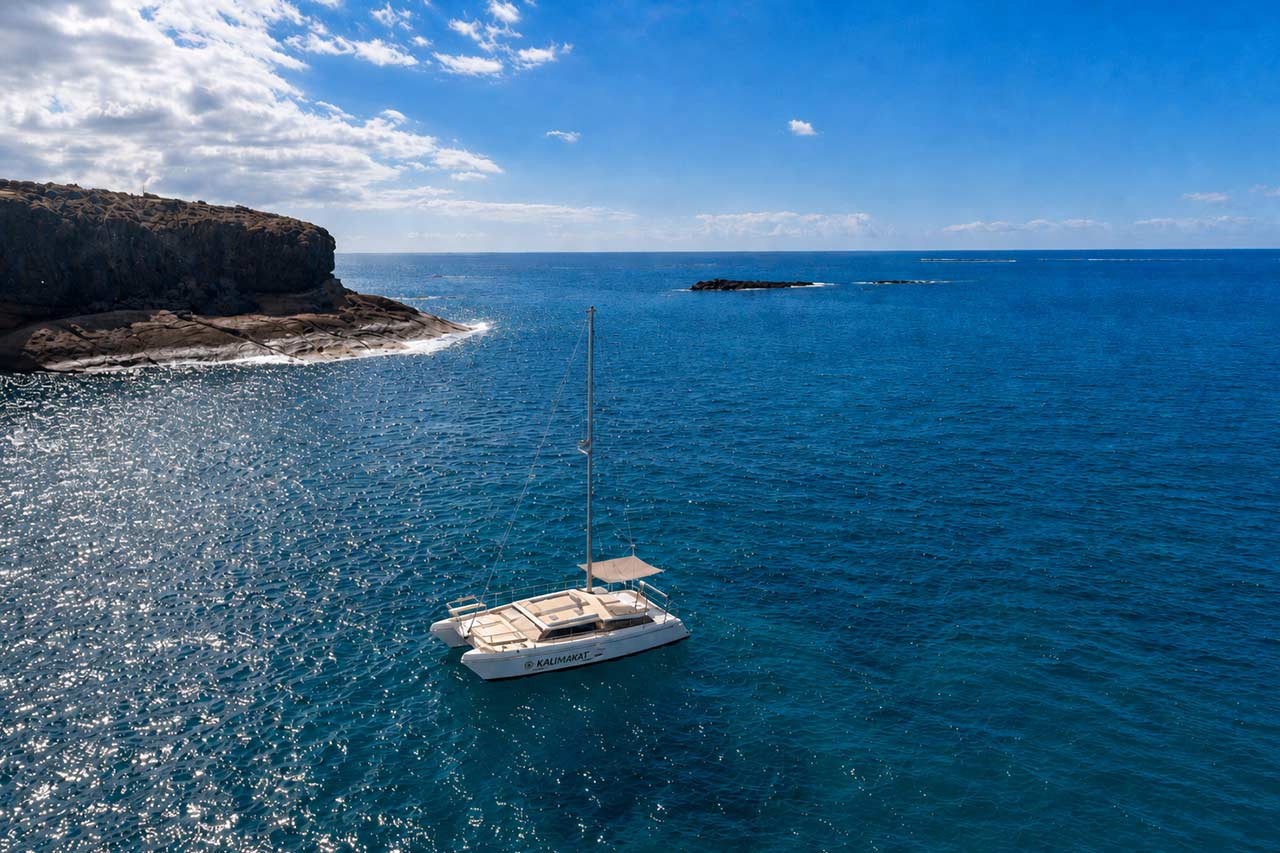 Luchtfoto van de witte catamaran Kalima Kat die op het diepblauwe Atlantische water vaart naast de rotsachtige kustlijn van Tenerife, zonder dat er gasten zichtbaar zijn.