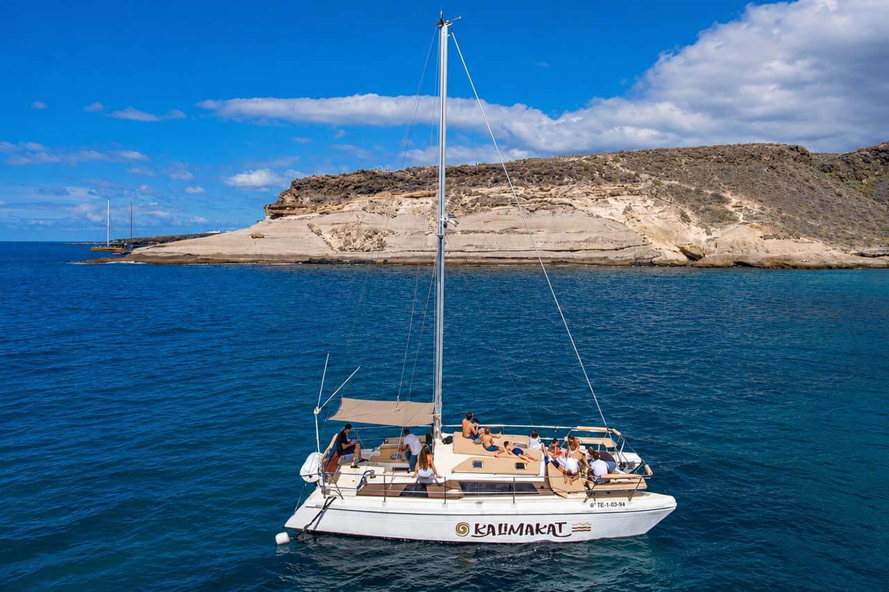 Kalima Kat catamaran met gasten die ontspannen op het dek in het blauwe Atlantische water naast de rotsachtige kustlijn van Tenerife.