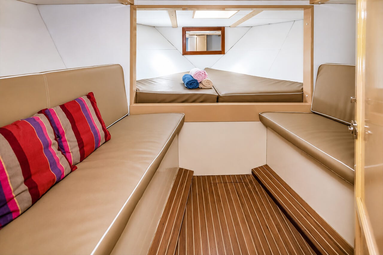 Neptuno’s clean lower cabin with cushioned seating, warm wooden flooring, colourful pillows, a forward resting area and neatly prepared towels.