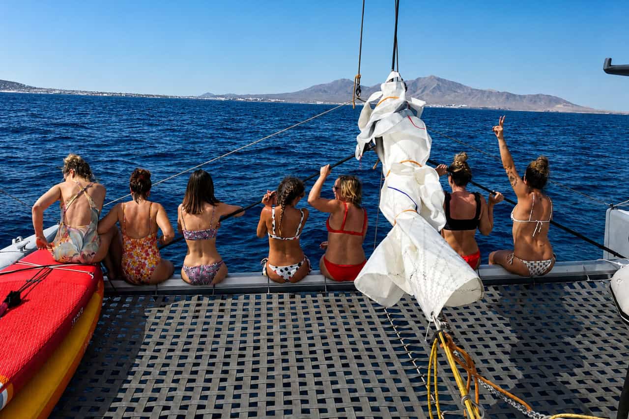 Group of women sitting together on the front net of a catamaran with ocean and volcanic mountains in the background