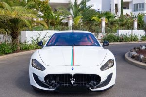 Een witte Maserati met een driekleurige streep (rood, wit en groen) op de motorkap geparkeerd op een straat. Op de achtergrond staan weelderige groene palmbomen en een wit hek rond woonhuizen.