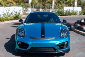 Een levendige turquoise Porsche 718 Cayman met een opvallende zwarte streep in het midden, geparkeerd op een zonovergoten oprit omringd door weelderige tropische tuinarchitectuur en witte privacy muren. De sportwagen heeft opvallende afgeronde koplampen en een gestroomlijnd, aerodynamisch ontwerp.