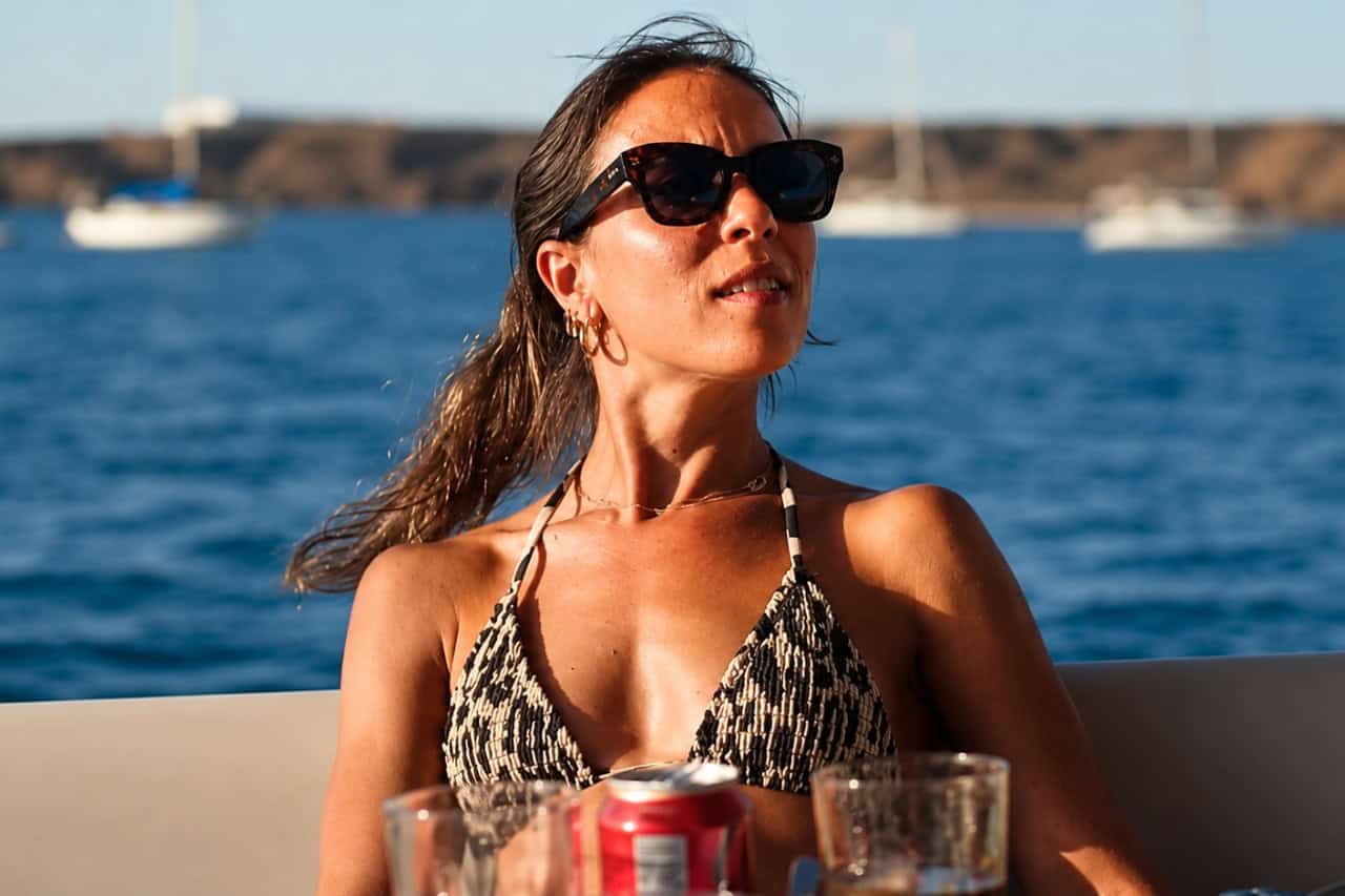 Woman wearing sunglasses relaxing on a private boat with the blue ocean and anchored sailing boats in the background