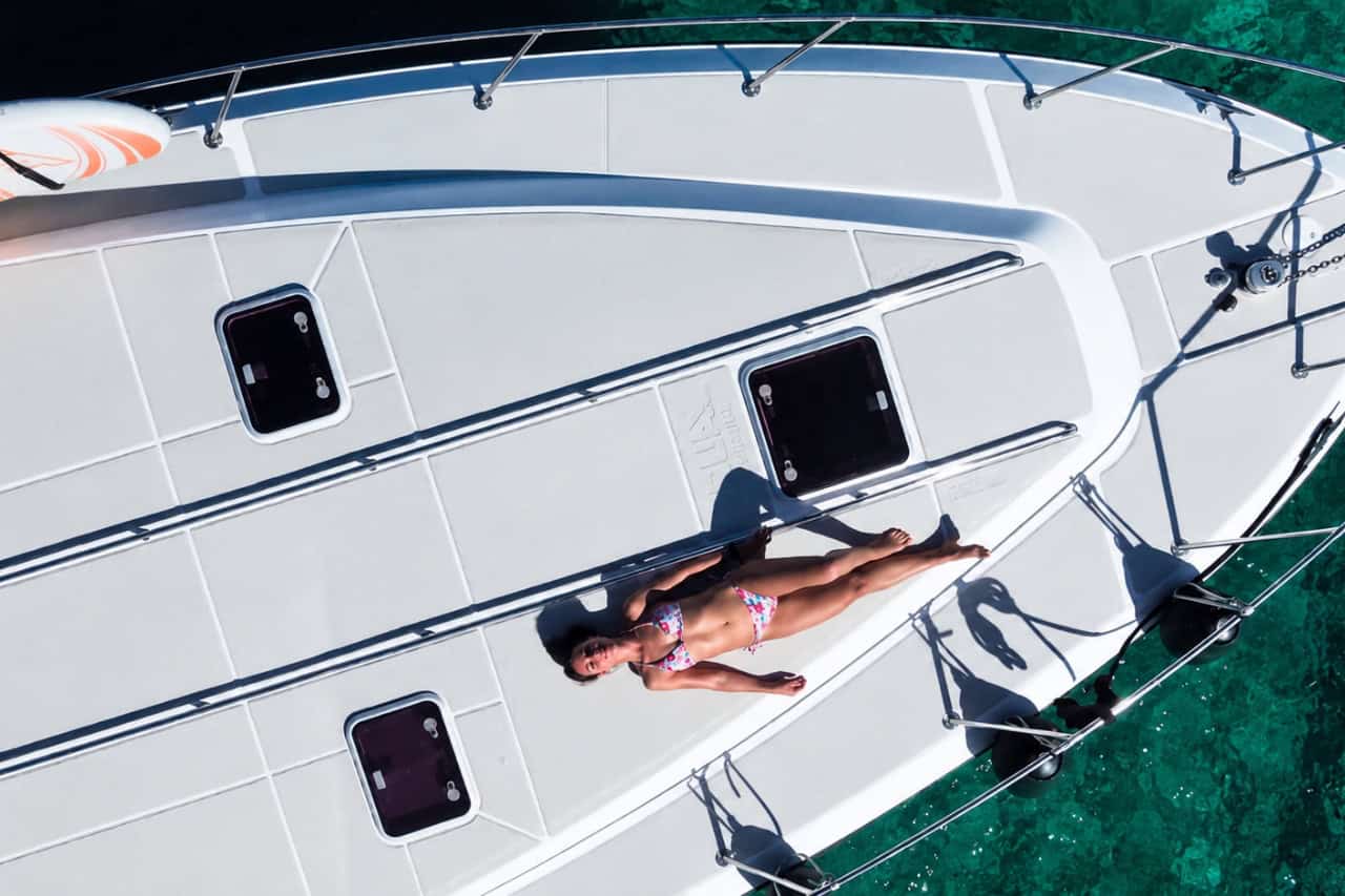 Aerial view of a woman sunbathing on the spacious white bow of a luxury yacht above clear turquoise water