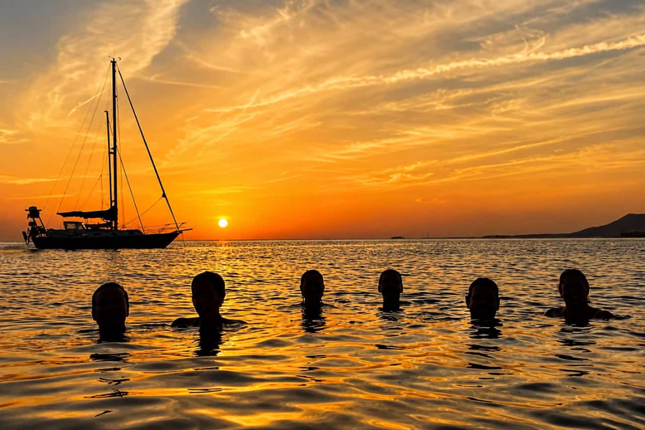 Group of people swimming in calm ocean water at sunset with a sailing yacht silhouetted on the horizon