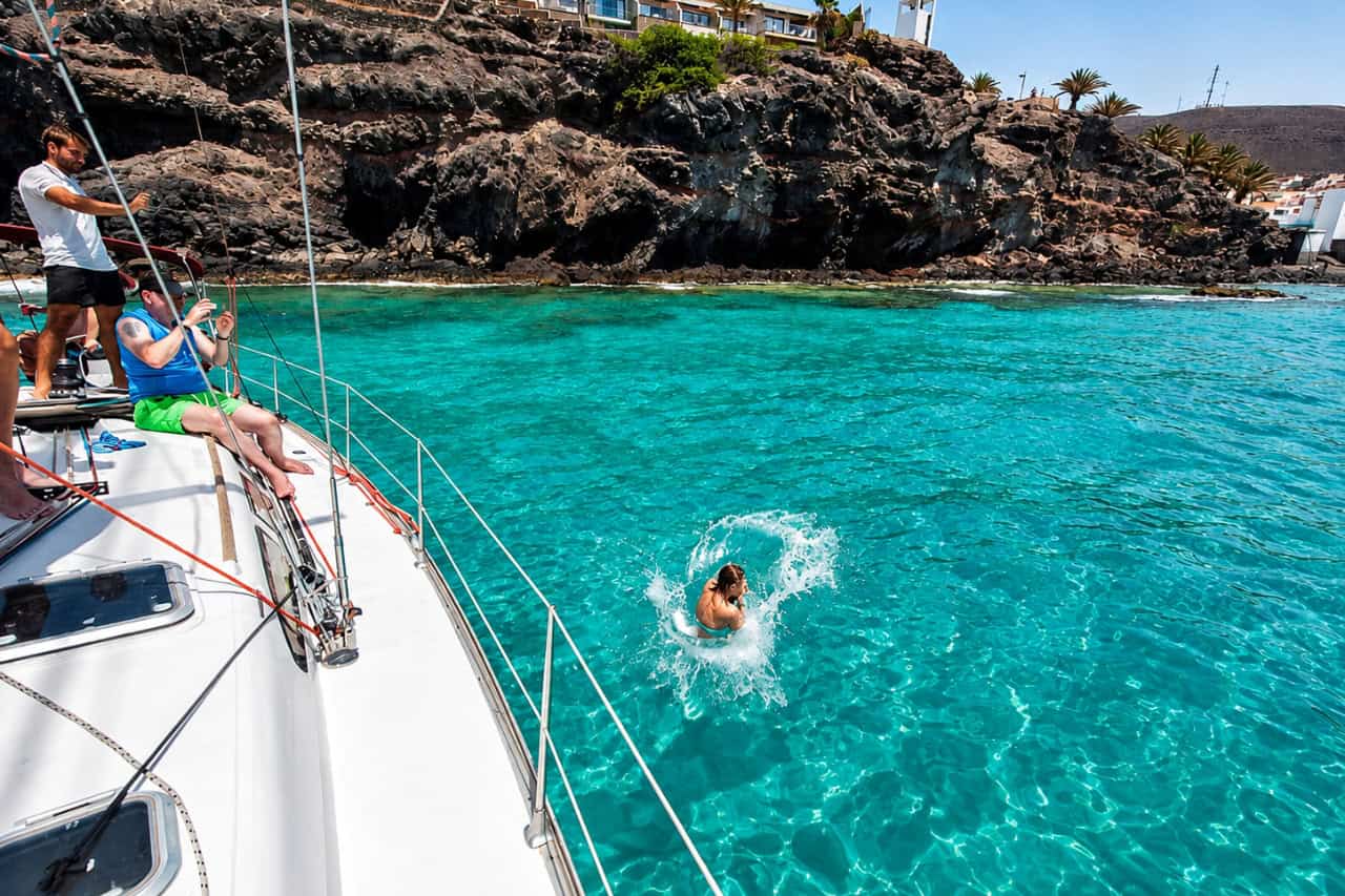 Guest jumping from a sailing yacht into clear turquoise water beside the volcanic coast of Fuerteventura
