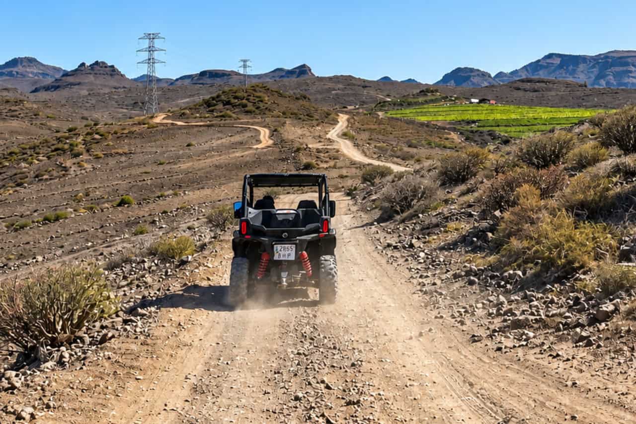 Buggy driving along a dusty off-road trail in Gran Canaria through open countryside with mountains, vegetation and green farmland ahead.