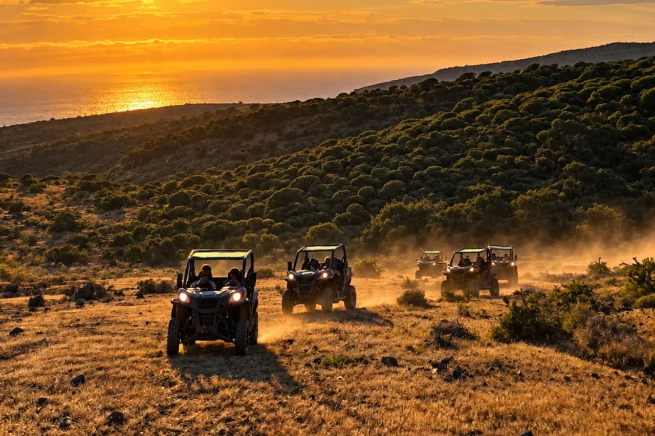 Buggy convoy driving across a dusty off-road landscape in Gran Canaria with mountain vegetation and the Atlantic Ocean in the background.
