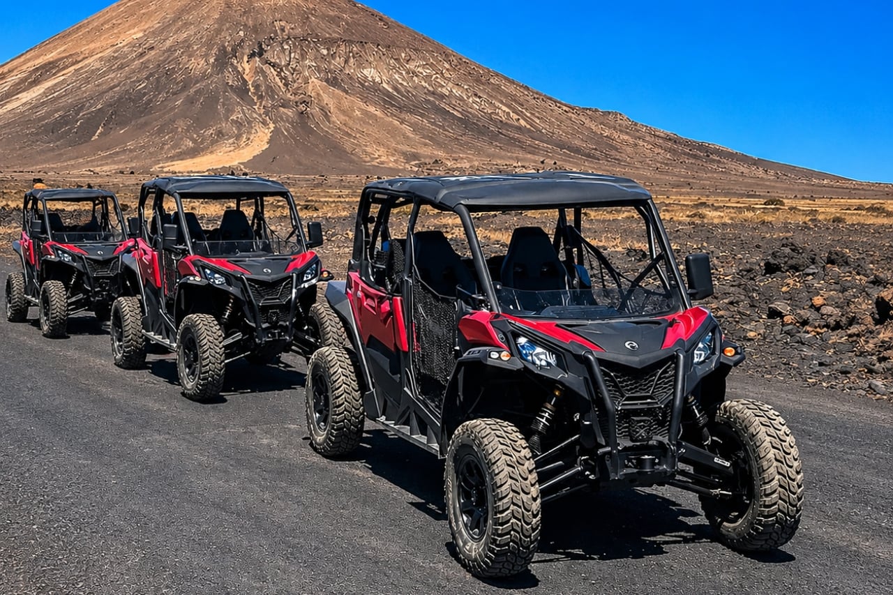 A convoy of three red and black off-road buggies positioned on a dark volcanic track, with a large mountain and clear blue sky in the background.