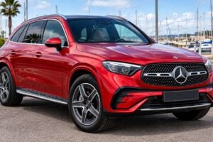 En röd Mercedes-Benz GLC 300 de parkerad bredvid en marina på Teneriffa. Bilen visas från fram-vänster vinkel, med en stor Mercedes-grill, LED-strålkastare, takrails i silver, lättmetallfälgar, tonade bakrutor, kromdetaljer och en glansig röd lack. Båtar, palmträd och marinan syns i bakgrunden.