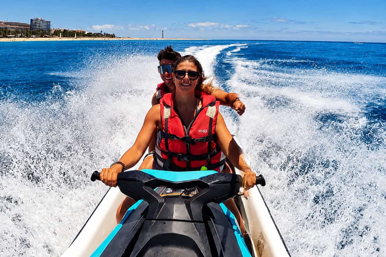 Smiling couple riding a jet ski with a trail of white spray near Morro Jable