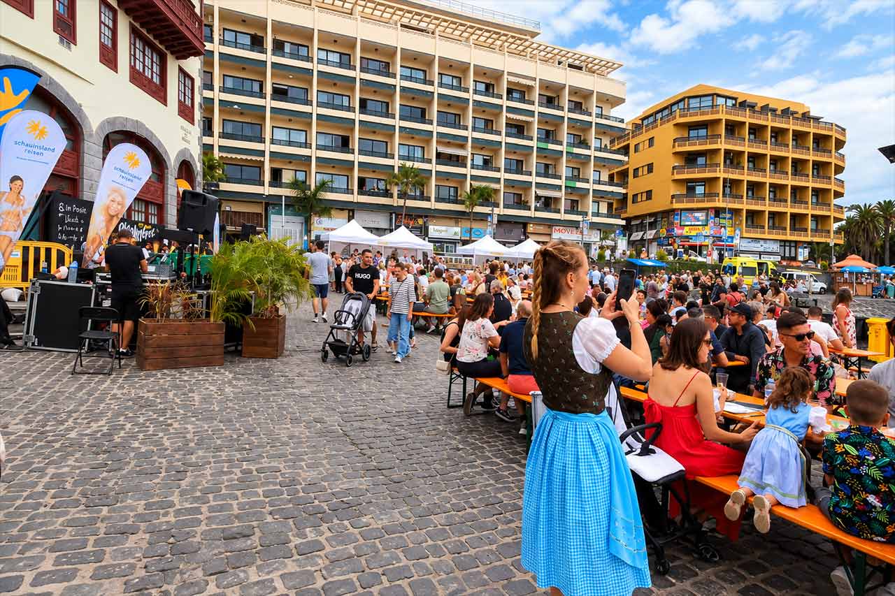 O mulțime animată pe timp de zi umple sărbătoarea Oktoberfest din Puerto de la Cruz, cu mese comune, tarabe de festival și atmosferă bavareză în apropierea frontului maritim din nordul insulei Tenerife.