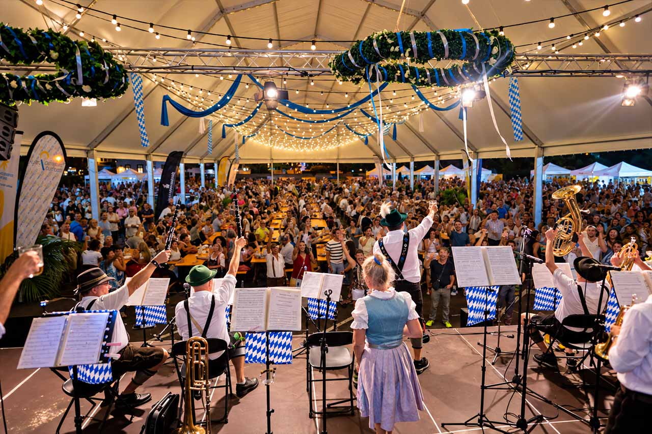 O orchestră bavareză cântă sub cortul decorat de la Oktoberfest, în timp ce oaspeții se adună în jurul meselor comune pentru muzică live de aramă, băuturi și o seară plină de energie.