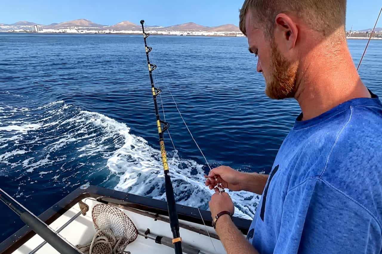 Man preparing fishing line and tackle aboard a boat with the coastline and volcanic hills of Lanzarote in the background.