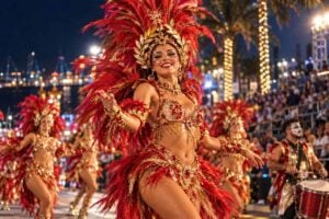 Impression of Ritmo y Armonía 2027 in Santa Cruz de Tenerife: a comparsa dancer in red and gold feathers with a laurel headdress on Avenida de Anaga at night, tribune and harbour lights behind