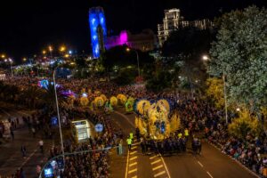 Panoramiczny nocny widok na Karnawał w Santa Cruz de Tenerife, z spektakularną złotą platformą paradną poruszającą się ulicami wypełnionymi widzami. Kolorowi performerzy, palmy, światła miasta i oświetlone budynki tworzą imponujący przegląd skali i atmosfery słynnych karnawałowych uroczystości na Teneryfie