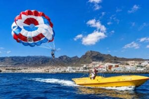 Varjohiihtoa Teneriffalla valtameren yllä lähellä Costa Adejea taustallaan keltainen pikavene ja vuoristoinen rannikko.