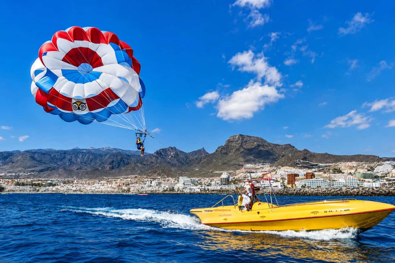 Parasailing in Tenerife over the ocean near Costa Adeje with a yellow speedboat and mountain coastline in the background.