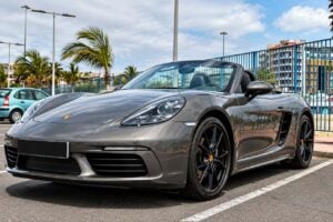 Metallgrå Porsche 718 Boxster cabriolet tillgänglig för uthyrning på Teneriffa, visad från främre trekvartsvinkeln med taket nere, svarta lättmetallfälgar, Porsche LED-strålkastare, svart läderinteriör och sportig låg profil i en solig kustmiljö.