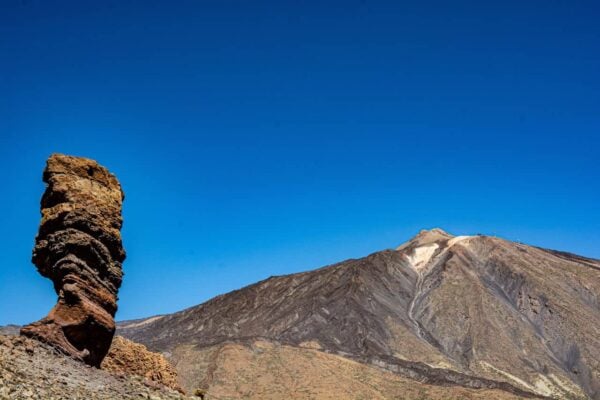 Levende landskab med den ikoniske Mount Teide under en klar blå himmel. I forgrunden står en markant formet, tårnhøj klippeformation tydeligt frem mod baggrunden af bjergets barske skråninger.
