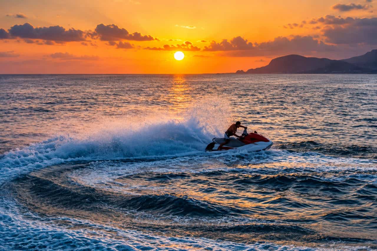 Jet ski rider carving through the ocean at sunset, with a glowing sky and mountainous coastline in the background.