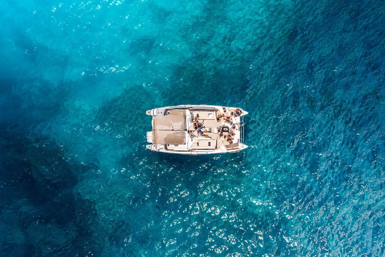 Top-down luchtfoto van de Kalima Kat catamaran drijvend op helder turquoise Atlantisch water bij Tenerife, met zicht op het ruime dek en de omliggende zee.