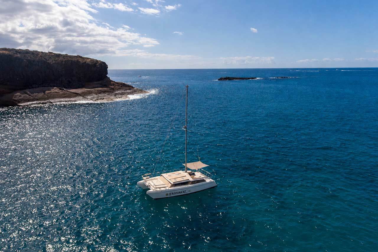 De catamaran Kalima Kat alleen op helderblauw Atlantisch water naast de rotsachtige kustlijn van Tenerife, weergegeven als een strakke, schilderachtige bootweergave zonder zichtbare gasten.
