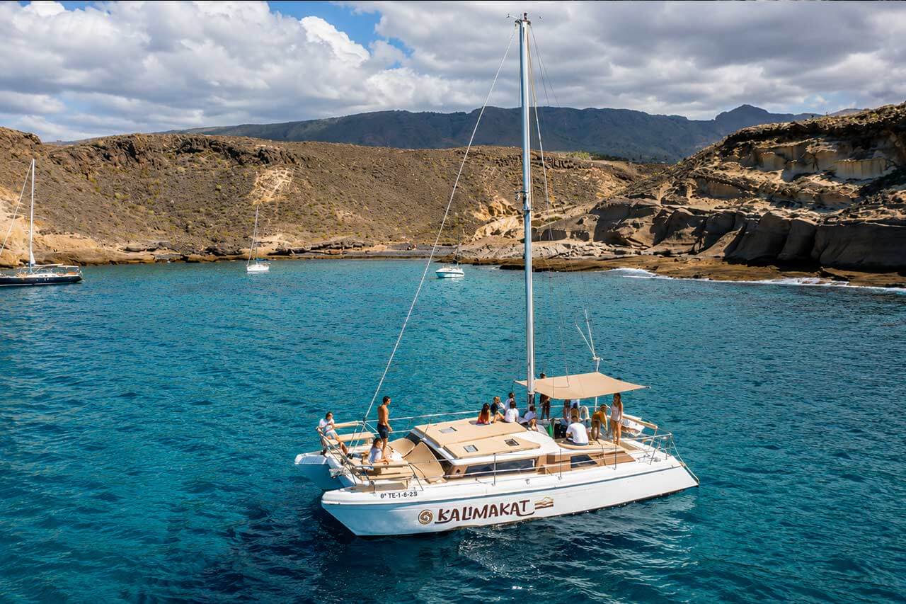 De Kalima Kat catamaran vaart langs de rotsachtige kustlijn van Tenerife met gasten die op het dek ontspannen, omringd door helderblauw Atlantisch water.
