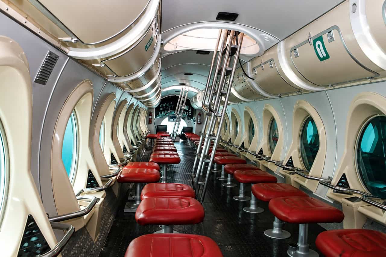 Interior of a tourist submarine with red seats and large windows for viewing marine life underwater.