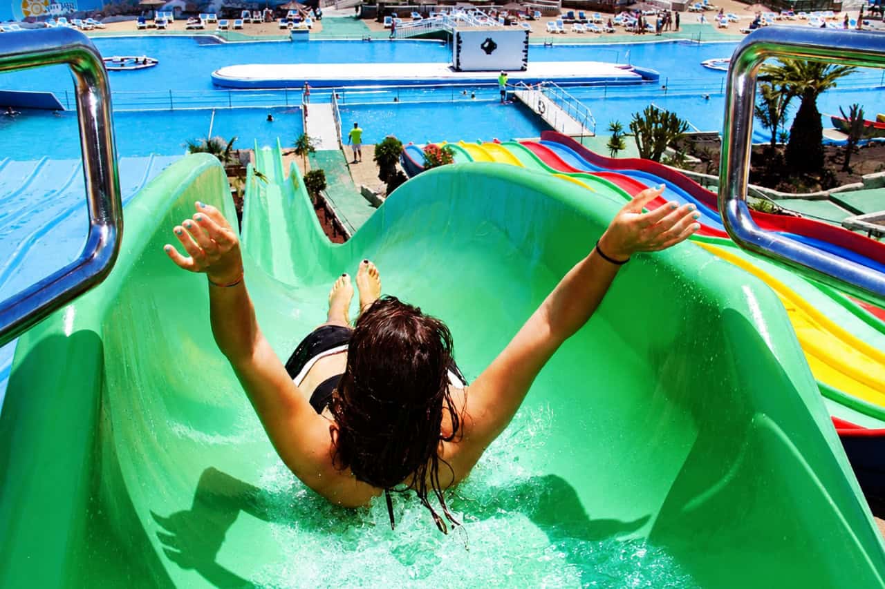 Visitor sliding down a green water slide with arms raised at a water park in Fuerteventura.
