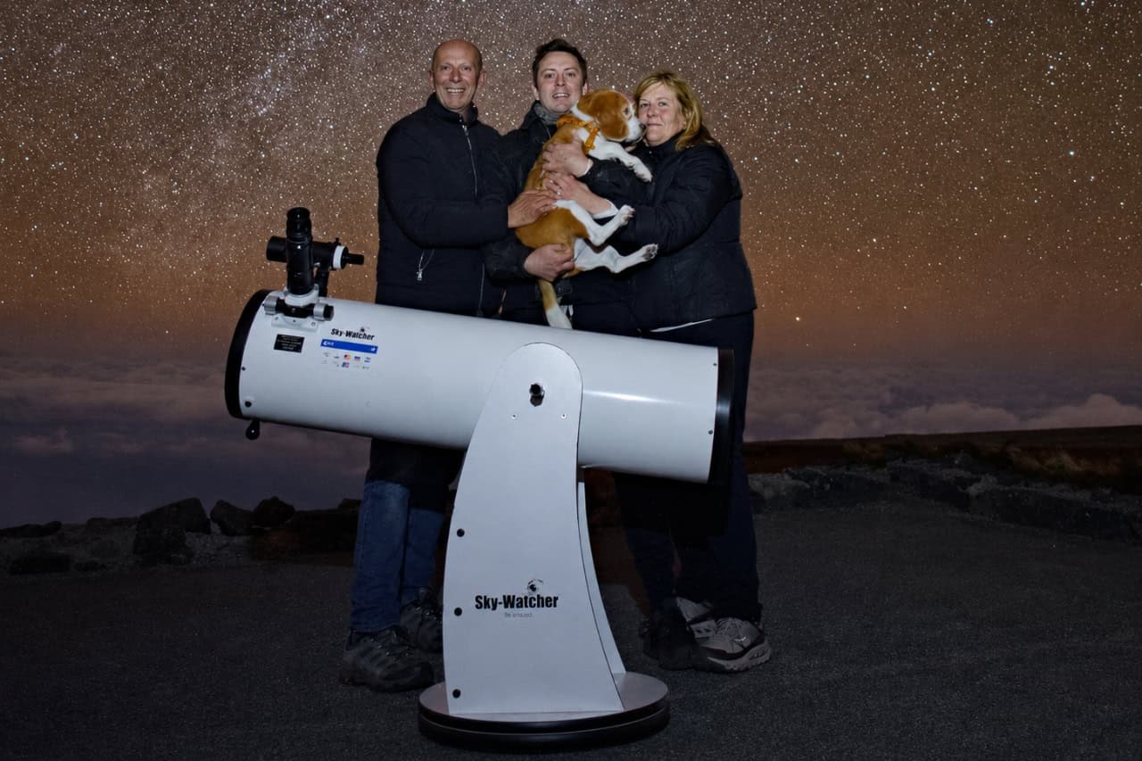 Three people holding a small dog beside a large telescope under a star-filled night sky in La Palma.