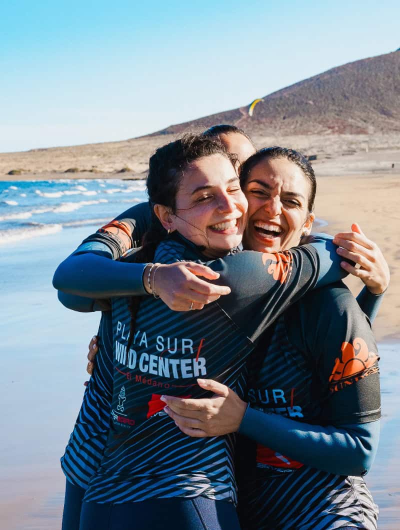 Try water sports Tenerife - friends laughing and hugging after a surf session at Playa Sur Wind Center El Médano booked with Club Canary