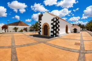 Traditional white church with black volcanic stone corners in Fuerteventura, surrounded by a spacious plaza with palm trees and volcanic mountains under a bright blue sky.