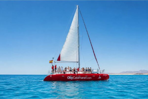 Large red sailing catamaran cruising off the coast of Fuerteventura during a private party boat charter from Corralejo with guests relaxing on deck.