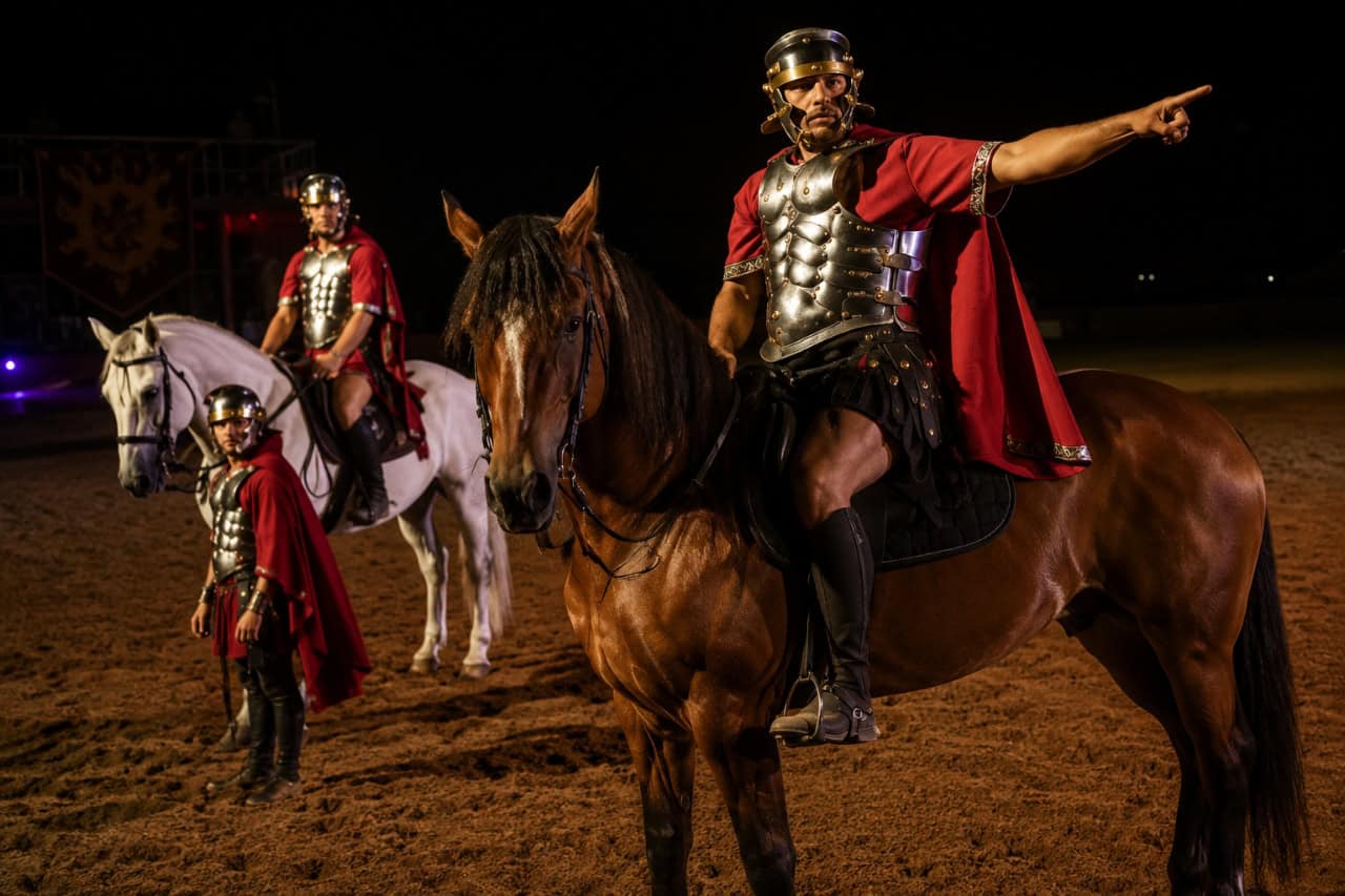 Gladiator performers in Roman armour riding horses during a live equestrian show at Hipica Xanadú in Tenerife under dramatic evening arena lighting.