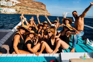 Happy group posing on the front of the Boat Party Gran Canaria catamaran with the volcanic coastline in the background.