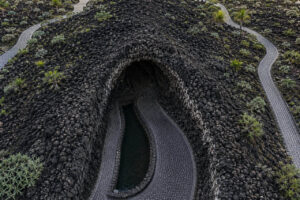Aerial photograph of the Jameos del Agua cave entrance in Lanzarote, featuring volcanic lava rock, a winding stone pathway, and a tranquil underground lagoon surrounded by the island's unique volcanic landscape.