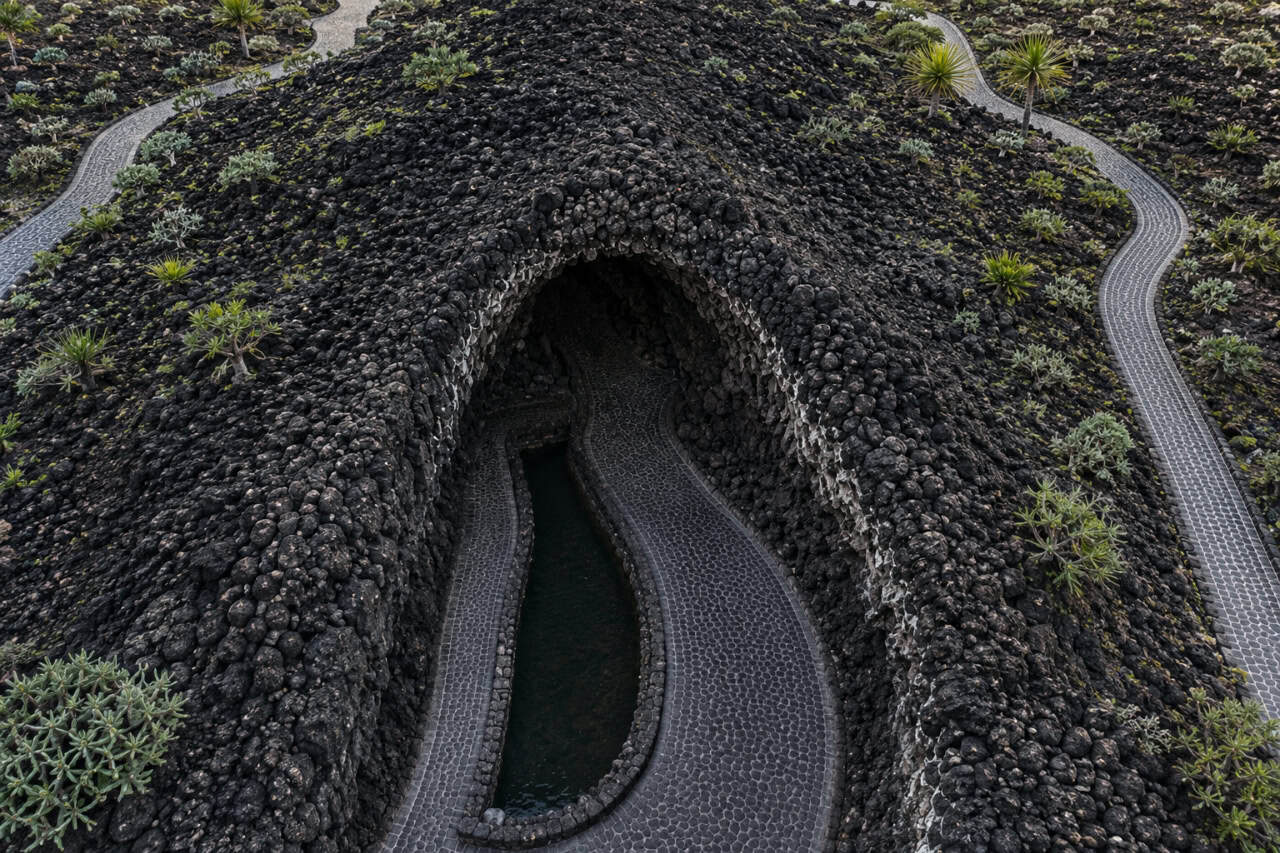 Aerial photograph of the Jameos del Agua cave entrance in Lanzarote, featuring volcanic lava rock, a winding stone pathway, and a tranquil underground lagoon surrounded by the island's unique volcanic landscape.