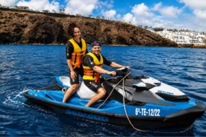 Smiling couple wearing life jackets on a Sea-Doo jet ski in the clear blue waters of Lanzarote, with volcanic cliffs and coastal resorts in the background.